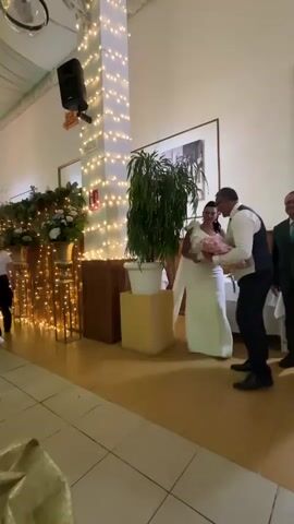 A wedding reception is underway. A man in a suit and a woman in a white wedding dress walk down an aisle adorned with string lights and greenery. The man carries a bouquet of pink flowers. Another man in a suit is visible in the background, holding a piece of white fabric. The room has tiled floors and a white ceiling with green drapes. Tables are set with white cloths and gold accents.