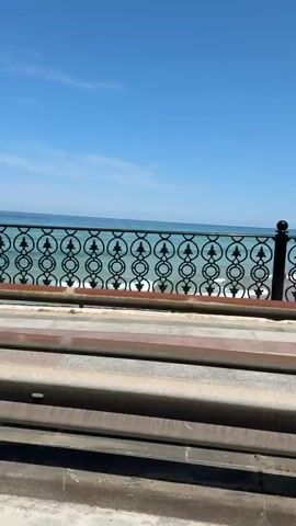 This is a first-person perspective video of a road next to a body of water. The camera is in motion, suggesting the video is being filmed from a moving vehicle. The foreground features a concrete road and a rusty metal guardrail. In the midground, there is a patterned walkway and a decorative black metal fence with circular motifs. Beyond the fence, the ocean stretches out under a clear blue sky. The water appears calm with small waves. The sun is shining, indicating it is daytime. No people or animals are visible in the frame. The overall scene depicts a scenic view of a coastal road.