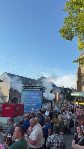 A large crowd of people are gathered outdoors, looking towards a stage. Confetti is being released from the stage, creating a smoky effect. There are buildings in the background and a clear blue sky overhead. The scene appears festive, likely a celebration or event.