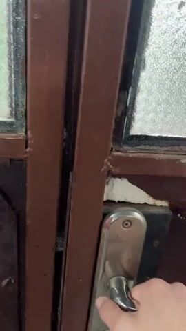 The video begins with a close-up of a brown door frame with glass panels. A hand is seen reaching for and turning a silver door handle. As the door opens, the camera moves outward to reveal a scene of heavy hail falling on a gravel path and a dirt area. In the background, there is a red, rusty trailer with a black door and a window, some trees, and a building. The ground is covered in a thick layer of hailstones. The hail appears to be small, like peas, and is falling densely. The environment suggests an outdoor setting, possibly a yard or a street. The weather is overcast, indicating a storm.