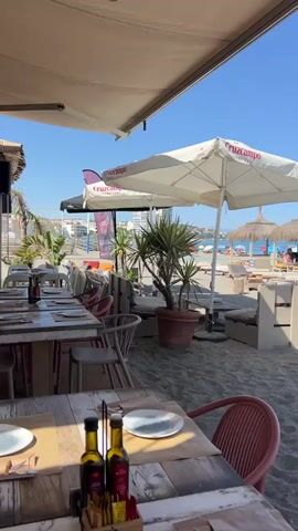 The video opens with a shot of tables and chairs set up on a sandy beach, suggesting an outdoor restaurant or beach club. In the foreground, a wooden table is set with plates, cutlery, and bottles of olive oil and balsamic vinegar. Behind it, a red chair is visible. A large white umbrella with a logo that reads "Cruzcampo" is prominently displayed, providing shade over a seating area. Palm trees and other potted plants are scattered around.

The camera pans to the right, revealing more of the beach scene. Several straw-roofed cabanas or beach huts are visible, furnished with lounge chairs and cushions. People are seated in some of these areas, relaxing and enjoying the sunny weather. Some individuals are sunbathing on loungers, while others appear to be socializing.

Further into the background, beach umbrellas, sunbeds, and a few people can be seen on the sand. The ocean is visible in the distance under a clear blue sky. A long wooden walkway extends from the foreground onto the sand. The overall atmosphere suggests a leisurely day at a popular beach destination, possibly a resort area.