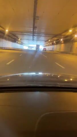 A car is driving through a tunnel. The tunnel has a concrete ceiling with metal fixtures. There are lights along the tunnel walls. The road is asphalt with white lane markings, including directional arrows. Other vehicles are visible further ahead in the tunnel. The view is from the driver's seat, focusing on the road ahead.