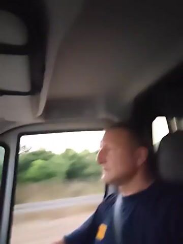 A man is driving a vehicle and looks at the camera and waves. The vehicle appears to be moving along a road with trees and foliage visible outside the window. The man is wearing a dark blue t-shirt with a yellow patch on the left side of his chest and a seatbelt. The interior of the vehicle shows part of the roof lining and a dark grey headrest.