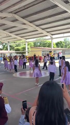 A group of children in matching lavender dresses are performing a dance routine in an outdoor courtyard. They are arranged in a circular formation on a gray, paved surface marked with red lines. The children are facing inwards towards the center of the circle and appear to be performing synchronized movements. In the background, adults and more children are observing the performance, some holding up cell phones to record. The courtyard is covered by a large, white, corrugated metal roof supported by exposed white beams, creating a shaded area. Beyond the covered area, a yellow building with red trim is visible, along with trees and a blue sky. Some children are wearing band uniforms on the far left of the frame. The perspective is from behind the audience, looking towards the performers.