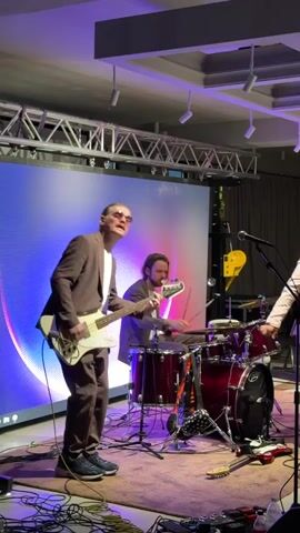 The video shows a band performing on a stage. In the foreground, a man in a brown suit and glasses plays a white bass guitar. He has long blonde hair pulled back in a ponytail. Behind him, a drummer in a grey suit plays a red drum kit. To the right, another person in a light pink suit with a pink bandana on their head sings into a microphone. A large LED screen with a colorful abstract design is visible behind the band. The stage is covered with a purple rug and there are many cables and audio equipment on the floor. The lighting is dim and colorful, suggesting an evening indoor event. The band appears to be playing a lively set, and the energy is palpable. The overall scene suggests a live music performance at an indoor venue, possibly a concert or a private event. There is no visible text in the video.