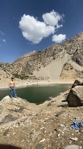 A man stands on a rock near a body of water. He is shirtless and wearing blue jeans. The man is looking towards the camera. The body of water is surrounded by mountains. The sky is blue with some white clouds.