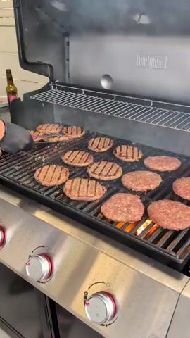 The video shows a close-up of a Weber grill outdoors, with numerous hamburger patties cooking on the grates. The patties are well-marked with grill lines, and some are beginning to flare up with flames. A black-gloved hand is visible in the upper left corner, holding a pair of tongs and appears to be adjusting or flipping the patties. Smoke rises from the grill, indicating active cooking. A bottle of beer is visible on a counter behind the grill. The scene suggests a casual outdoor gathering or barbecue. The setting appears to be a backyard or patio area, likely in a sunny environment given the brightness of the scene. The time of day is likely daytime, based on the lighting. The visible text on the grill is "WEBER". The location context is Lod, Israel.