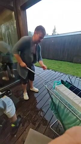 A person is mopping a wet wooden deck. The person is wearing a grey sweater, black shorts, and white Crocs. They are holding a mop with a blue and white mop head. There is a drying rack with clothes on it to the right of the person. Another person's legs are visible in the lower left corner of the frame. The deck appears to be outdoors, possibly on a porch or patio, with a wooden fence and grass in the background. The lighting suggests it may be daytime, but overcast.