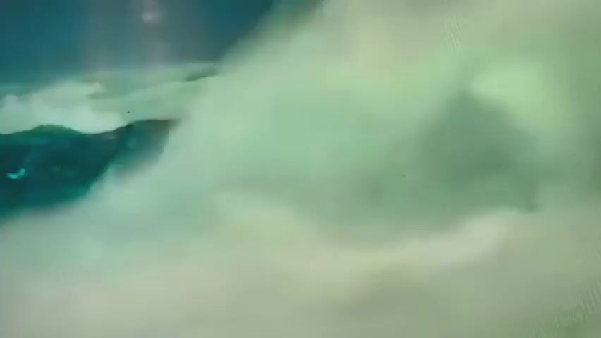 The video shows a dark, rocky formation emerging from churning, light-colored mist or water. The formation appears to be large and rugged, with various shapes and sizes of rocks clustered together. White mist or spray is actively obscuring parts of the rocks, suggesting movement or a turbulent environment. The background is a hazy, muted green, contributing to a sense of mystery or dramatic atmosphere. The lighting is soft and diffused, with no distinct light sources visible. The overall impression is one of a powerful natural force at play, with the rocky mass battling against the elements.