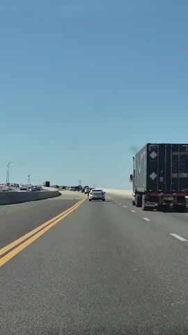The video shows a view from a vehicle traveling on a multi-lane highway. The sky is clear and blue. On the right side of the highway, a large semi-truck with a black trailer carrying what appears to be an Amazon logo is traveling in the same direction. In the distance, on the left side of the highway, there is a line of cars, suggesting traffic congestion. The highway is paved with visible lane markings. The perspective is from the driver's seat or a similar vantage point within a vehicle.