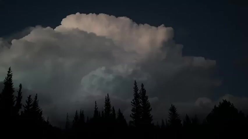 The video shows a time-lapse of a large, dark cloud formation against a deep blue night sky. The cloud is illuminated from within by a bright light, suggesting lightning. In the foreground, a silhouette of a dense forest of pine trees runs along the bottom of the frame. The trees are dark and indistinct, their tops reaching up towards the imposing cloud. The video captures the dynamic movement of the cloud, with sections of it appearing to swell and shift.