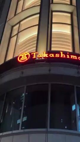 The video shows a low-angle, upward shot of a building at night. The building is identified as "Takashimaya S.C." by a lit-up red sign. The architecture of the building is modern, with curved windows and illuminated vertical panels. The top portion of the building shows some interior lighting through the windows. The background is a dark sky. The camera slowly pans upwards, following the height of the building.