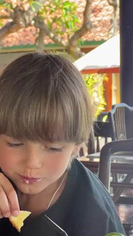 A close-up video focuses on a young boy with brown hair. A small green caterpillar is perched on top of his head, near the crown. The boy is looking to his left, with his mouth slightly open as if he is eating. The background is blurred, but appears to be an outdoor setting with trees and structures that suggest an Asian architectural style. The lighting is bright, casting a warm glow on the scene.