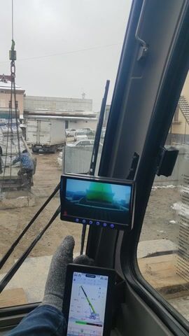 The video shows a view from inside the cab of a crane. The crane is lifting a load of bagged materials from the back of a truck. Two people are visible on the truck, helping to secure the load. The crane operator appears to be using a tablet and a screen mounted inside the cab to assist with the lifting operation. The environment appears to be an industrial area with storage containers and other buildings in the background. The weather is overcast and there is some snow on the ground.