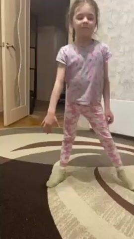 A young girl demonstrates a split. She wears pink patterned pajamas and green socks, and her hair is in two braids. She starts in a wide stance with her hands on the rug. She slowly lowers herself to the floor, stretching her legs into a split. She looks up at the camera and says something, then lowers her upper body completely to the floor. The girl performs the split on a round rug with a brown and beige pattern. In the background, a door is slightly open revealing a room with various items. A radiator is mounted on the wall. The walls have a wallpaper with a subtle pattern.