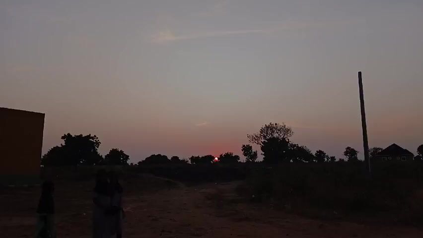 Two girls in school uniforms walk away from the camera along a dirt road. A building stands on the left side of the frame and a tall pole on the right. In the background, silhouetted trees and a house stand against the horizon. The sun is setting, appearing as a red glow through the trees. The sky is a mix of gray and pink hues. The environment suggests a rural setting.