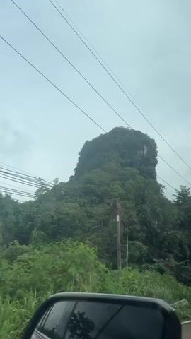 This media file captures a view from a moving vehicle, likely a car, in Ao Luek, Thailand. The scene is dominated by a lush, green, and imposing limestone karst mountain that appears to be covered in dense vegetation. The mountain rises steeply, creating a dramatic backdrop against a cloudy, overcast sky.

The foreground shows parts of the vehicle, including a side mirror and the windshield with a sticker attached. This sticker, visible at 00:01, appears to be a vehicle registration or inspection sticker, featuring some text and circular emblems, though the specific details are difficult to discern due to the blur and angle.

The environment suggests a tropical or subtropical climate, with the abundant greenery and the overcast weather. The presence of power lines and poles running alongside the road indicates that this is a developed area, likely a highway or main road. The road itself is visible, with other vehicles appearing in the distance, suggesting ongoing traffic.

The overall mood is one of travel and natural beauty. The cloudy weather might contribute to a slightly subdued atmosphere, but the sheer scale and verdant appearance of the karst formation are undeniably impressive. The time of day is not precisely indicated, but the overcast conditions suggest it could be daytime. The location is confirmed as Ao Luek, Thailand, known for its distinctive karst landscapes.