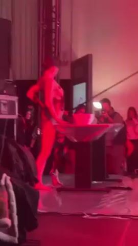 A performer in a dark, floral-patterned bodysuit stands barefoot on a lit stage. To the left, a sound system is visible, and a table covered in clear plastic stands center stage. A large white bowl rests on the table. Behind the table, a screen displays white text. The performer motions with her arms and adjusts her stance. Several spectators, some holding cameras, are visible in the background. The lighting is predominantly red, giving the scene a vibrant hue.