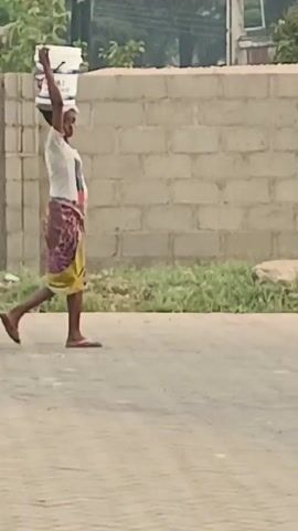A woman is walking along a paved road while carrying a white bucket on her head. She is wearing a white t-shirt, a patterned wrap skirt, and flip-flops. She is walking away from the camera. Behind her is a brick wall and some greenery, including what appears to be a palm tree. There is also a water tank visible in the background. The lighting suggests it is daytime.