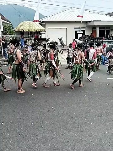 A group of people in traditional attire are marching down a street. The individuals are wearing body paint, grass skirts, and some are carrying spears. They appear to be participating in a parade or cultural event. In the background, there are buildings, trees, and spectators watching the procession. The sky is overcast, suggesting a cloudy day. The overall scene depicts a cultural celebration taking place in an outdoor, possibly rural, setting.