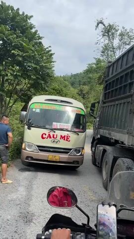The video shows a narrow road blocked by a small white and beige bus facing the camera and a large, dark-colored truck. The bus displays "HÀ GIANG - YÊM MINH" and "CÂU MÈ" in bold red letters. Its license plate reads "23F 000.86." A man wearing a blue shirt and dark shorts walks towards the bus from the left, while another man in a darker blue shirt stands near the front of the truck. The video is recorded from the perspective of someone on a motorcycle, with the motorcycle's handlebars, mirror, and a phone mounted on the handlebars visible. The phone screen is active. Lush green trees and hills surround the road. The sky is overcast. The scene appears to be a traffic obstruction or impasse on a rural, winding road.