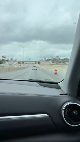 A driver's point of view shows traffic on a highway on an overcast day. The road has orange and white traffic barrels and a white SUV is ahead of the car. A sign indicates the road ahead goes West to Loop 1604.