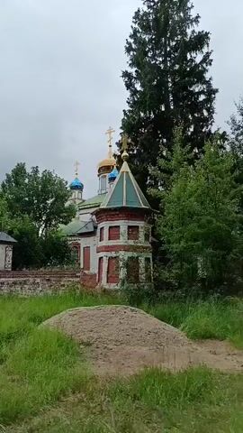 A drone shot pans across a religious complex. The complex is made of brick and painted white with red trim. It features multiple domed structures topped with crosses, including one large golden dome. The complex is surrounded by a low brick wall with decorative pillars. Lush green grass and trees surround the structure, and the sky is overcast.