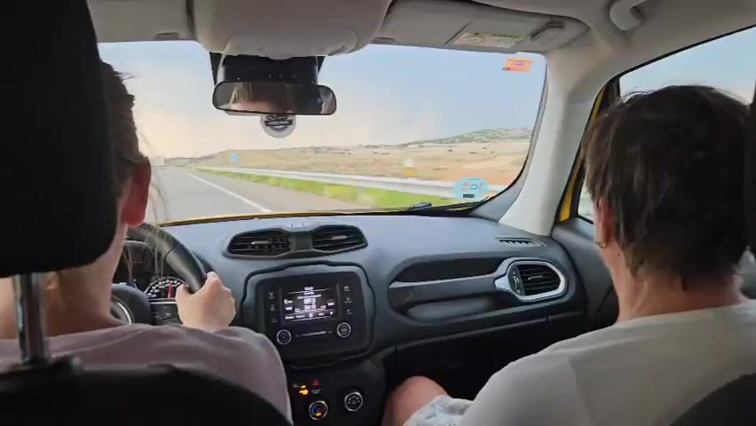 The video shows the interior of a yellow car from the perspective of the back seat. A woman with brown hair is driving, her hands on the steering wheel. A person with brown hair is in the passenger seat. The dashboard has a media screen and various controls. Outside the car, a highway stretches ahead, with a dry, hilly landscape on either side. The sky is overcast. The car appears to be moving.