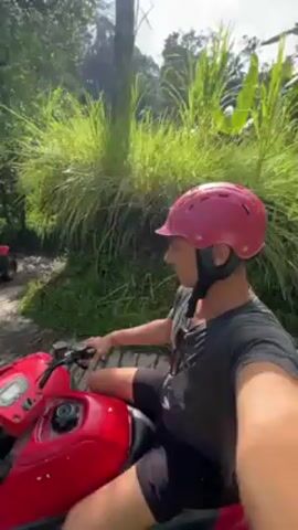 A man wearing a pink helmet and a gray shirt is riding a red ATV on a dirt path through a jungle-like environment. He is taking a selfie video. Two other ATVs are visible ahead of him on the path, each with a rider. Lush green vegetation surrounds the path. The weather appears bright and sunny.