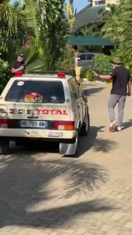 The video shows the rear of a vintage rally car parked on a brick-paved area surrounded by tropical vegetation. The car, painted in an off-white color, features racing decals including "TOTAL" prominently displayed. The license plate reads "HD-701-BN". Unusual, large mud flaps are attached to the rear wheels. Three people are visible. One man in a black shirt stands near the car observing, while another man wearing a black shirt and a hat walks away from the car. A woman wearing a green dress briefly walks past, taking a picture with her phone. The environment includes palm trees, flowering plants, and a building with a green roof in the background, indicating a tropical setting. The video appears to be taken on a sunny day, as evidenced by the strong shadows.