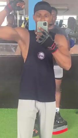A man in a black tank top and gray shorts takes a mirror selfie in a gym, flexing his bicep. He wears black fingerless gloves. Another man stands behind him in the reflection of the mirror. The gym has weight machines and a blue pillar.