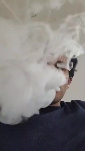 A person exhales a large plume of white vapor. The vapor surrounds their head and obscures most of their face. The person is wearing a dark blue sweater. The background is a white ceiling with a circular light fixture.