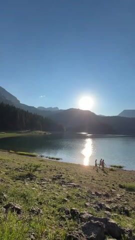 A group of people are walking along the shore of a lake. The sun is setting, casting a warm glow over the scene. The lake is surrounded by mountains and trees. The ground is covered in grass and rocks. One man is wearing a black t-shirt and shorts and is holding a phone. Another man is wearing a blue t-shirt and shorts and is holding hiking poles. A woman with blonde hair is holding a phone.
