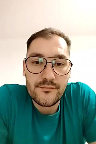 The video is a headshot of a man with short brown hair, a beard, and glasses. He is wearing a teal t-shirt. The background is a plain white wall. In the first two frames, the man looks directly at the camera. In the third frame, he touches his forehead with his hand and looks down slightly. In the fourth frame, he returns to looking at the camera.