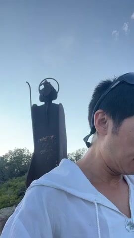 A man with short black hair and sunglasses on his head is taking a selfie. He is wearing a white hoodie. Behind him, there is a dark silhouette of a figure with a halo and a staff, possibly a statue or a sculpture. The background appears to be an outdoor setting with trees and a cloudy sky. The man is looking at the camera and speaking.