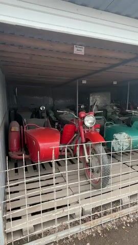 The video shows a collection of vintage motorcycles with sidecars stored in a covered outdoor area, possibly a shed or a long garage. The motorcycles are parked close together and appear to be mostly in red and green colors. The foreground features a red motorcycle with a sidecar, partially obscured by a metal grate fence. The motorcycles are parked on wooden pallets, which are stacked to create a slightly raised platform. In the background, other motorcycles with sidecars are visible, lined up in rows. Some appear to be in better condition than others, with some showing signs of wear and tear. Signs with text in what appears to be Cyrillic alphabet are attached to the wooden beams of the structure above. The lighting is somewhat dim, suggesting an overcast day or shade. The overall impression is that of a storage facility for antique vehicles.