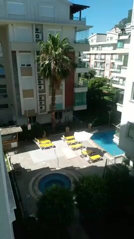 The video shows an aerial view of an apartment complex courtyard on a sunny day. The courtyard features a swimming pool, a circular hot tub, lounge chairs, a palm tree, and surrounding buildings. The camera pans from left to right, revealing the various elements of the courtyard. There are no people visible in the scene.