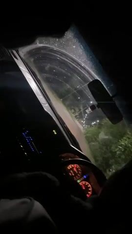The video begins with a close-up view of a car's dashboard at night. The speedometer needle is illuminated in red, indicating the car is in motion. The steering wheel is partially visible in the foreground, held by a gloved hand. Through the windshield, a dark, tree-lined street is visible, illuminated by distant streetlights. The overall impression is that of someone driving at night.