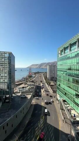 The video pans across a city by the sea on a sunny day. In the foreground, two modern buildings with glass facades dominate the view. One building, on the left, is mostly dark glass, while the building on the right features green glass. Between them, a road runs parallel to the coastline, with several cars and a van visible. Beyond the road, the sea stretches out, with a few boats dotted on the water. A large, older building with many balconies stands tall on the right side of the frame, reflecting the blue sky and surrounding buildings. Further in the distance, a mountainous coastline can be seen under a clear blue sky. The video captures an urban coastal landscape with a mix of modern and older architecture.