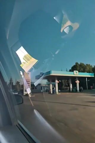 The video shows a view through a car window. The car is driving past a gas station. The gas station has a blue and white canopy and a sign with fuel prices. There are people walking around the gas station. The car continues driving, passing by trees and streetlights. The sky is blue.