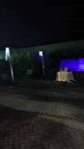 The video shows an outdoor event at night. In the foreground, a paved area with brick-like patterns is visible. In the mid-ground, there is a table covered with a white tablecloth, decorated with lights and what appears to be a cake. Behind this table, the scene is illuminated by purple lighting. Further back, a group of people are seen socializing under a covered area. The overall atmosphere suggests a celebration or gathering.