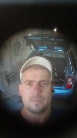 The video is a close-up selfie taken by a man wearing a white baseball cap and a dark t-shirt. He appears to be in a garage or workshop, with a blue car with its trunk open in the background. The environment is cluttered with tools and various objects. The lighting is dim, with some lights visible overhead. The man looks directly at the camera with a neutral expression. The video is shot from a low angle, looking up at the man.