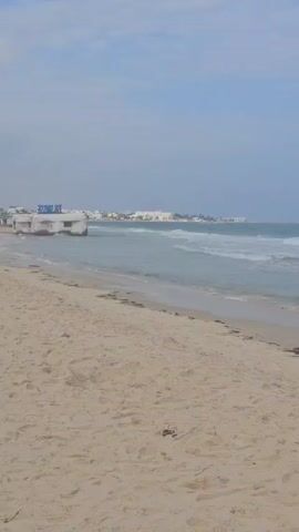 The video depicts a sandy beach meeting the ocean under a blue sky with scattered clouds. Gentle waves are breaking on the shore. There are some small pieces of seaweed or debris scattered along the shoreline. In the distance, there is a vague outline of buildings. The camera is stationary, capturing the scene from a wide angle. No people are visible in the frame.