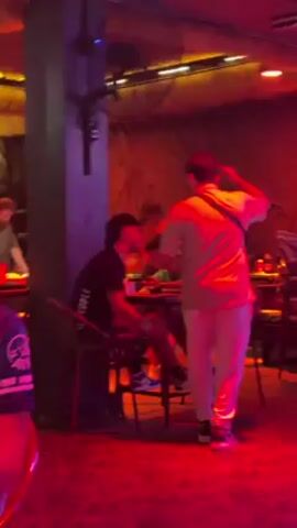 Three men are seated at a square table in a bar, illuminated by blue and red lighting. The man standing is wearing white pants and a white polo shirt with a black cross-body bag. He aggressively lifts a chair over his head, then slams it down on the ground, shattering it. Another man in a black t-shirt and patterned shorts watches from his chair. The third man in a green shirt remains seated at the table. The bar's interior features a textured wall, ceiling fans, and stacked chairs in the background.