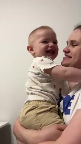 The video shows a mother holding her baby. The baby is wearing a white t-shirt with a pattern of blue cars and tan shorts. The mother is wearing a white t-shirt with a blue logo on the front. The baby is laughing and looking towards the camera. The mother is also smiling and appears happy. In the background, a second child, a boy, is visible. He is wearing a white t-shirt and is leaning in towards the baby. The overall scene is intimate and shows a moment of family interaction. The lighting suggests it is daytime, and the plain white wall in the background indicates an indoor setting, possibly a home. The emotions displayed are joy and affection. No weather or specific location cues beyond Germany are discernible from the visual information alone.