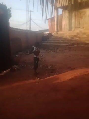 A young boy stands in a dirt yard, wearing a white tank top and dark shorts. He is holding something in his hands and appears to be making gestures with them. There are stairs visible in the background to the left, and a reddish-brown wall to the right. The ground is covered in dirt, debris, and a few plastic bottles. The lighting suggests it is daytime.
