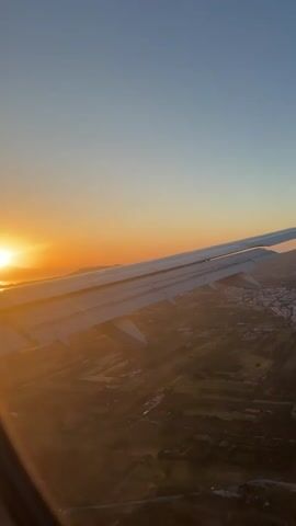 The video shows a plane wing against a sunset, with the ground visible below. The plane is flying over a landscape of fields and a small city, which appears to be in Europe, with a mountain range in the distance. The sun is setting on the horizon, casting a warm glow over the scene. The sky is clear and blue.