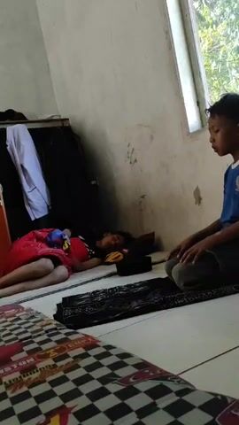 Two young children are in a room. One child is lying down asleep on the floor, covered by a red blanket. The second child is kneeling on a prayer mat, performing Islamic prayer. The room appears to be a simple living space with a window and a closet.