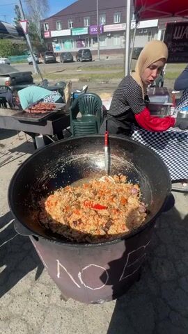 The video shows a food vendor at an outdoor market, preparing and serving pilaf. The vendor, a woman wearing a hijab, stands beside an enormous, black, cauldron-like pot filled with pilaf, a rice dish with meat, carrots, and red peppers. The pot is sitting on a base with the word "PLOV" painted on the side. She is wearing black gloves and a red sleeve over her arm. Behind the vendor, another person is grilling meat, and there is a red canopy, as well as other food preparation and serving areas. Cars are visible in the background, indicating the market is located near a street. The camera zooms in to provide a closer view of the pilaf in the pot. A large metal spoon is visible in the pilaf. A person off-camera asks someone to pass by.