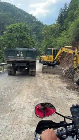 A person on a motorcycle is facing a large green truck with the license plate "12 H 002 82" and a yellow Hyundai excavator. The person is wearing a red shirt and a black helmet with a camouflage pattern. The motorcycle has a smartphone mounted on the handlebars, displaying a map. The road is partially paved and appears to be under construction. The environment is mountainous and lush with green trees. The excavator is positioned on a concrete pad, and the truck is waiting on the road.